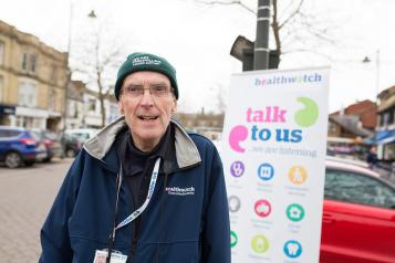 Older volunteer standing infront of a Healthwatch banner at an event